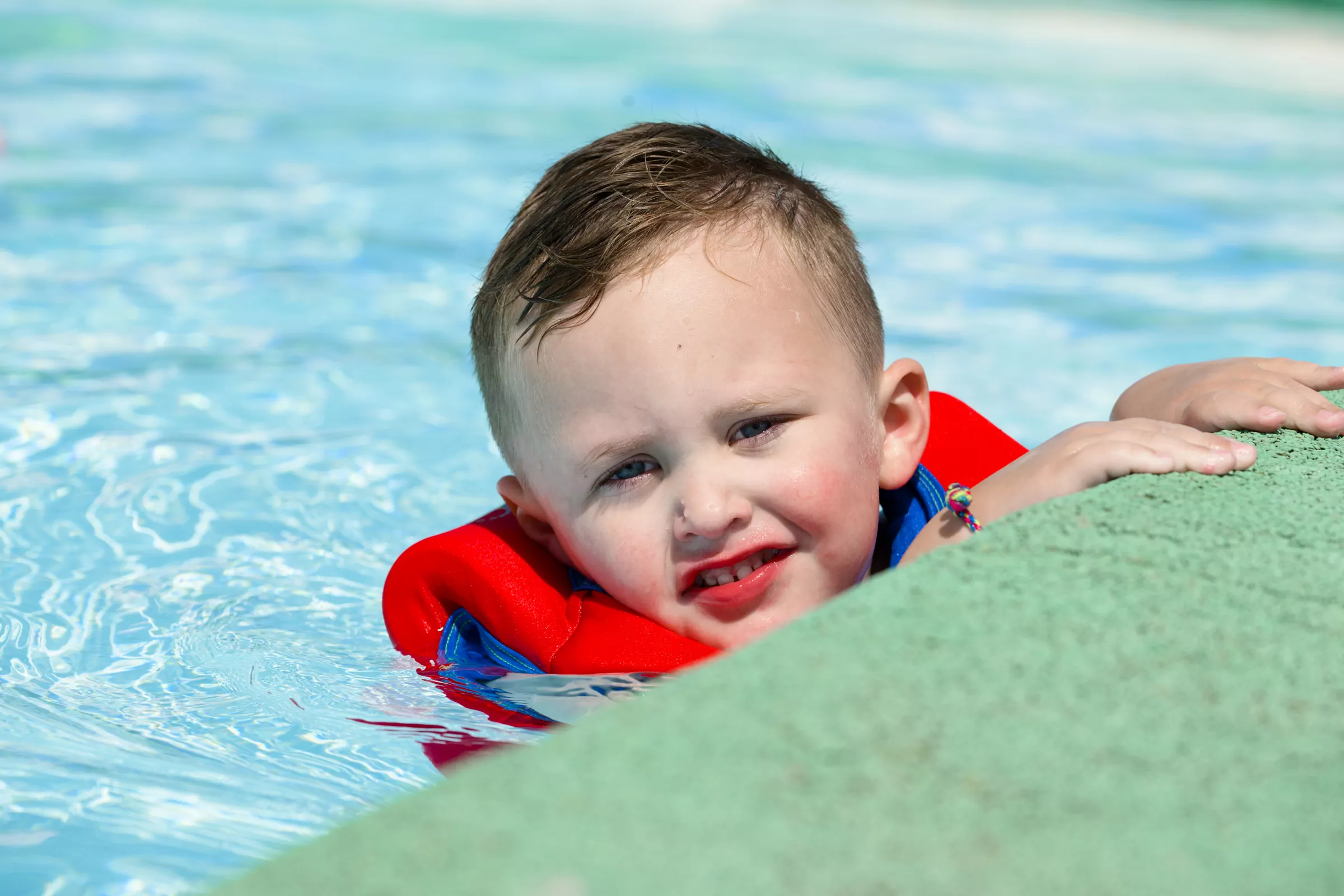 Young boy wearing a red life jacket enjoying swim time in the outdoor pool at Meadow Bay Villages.