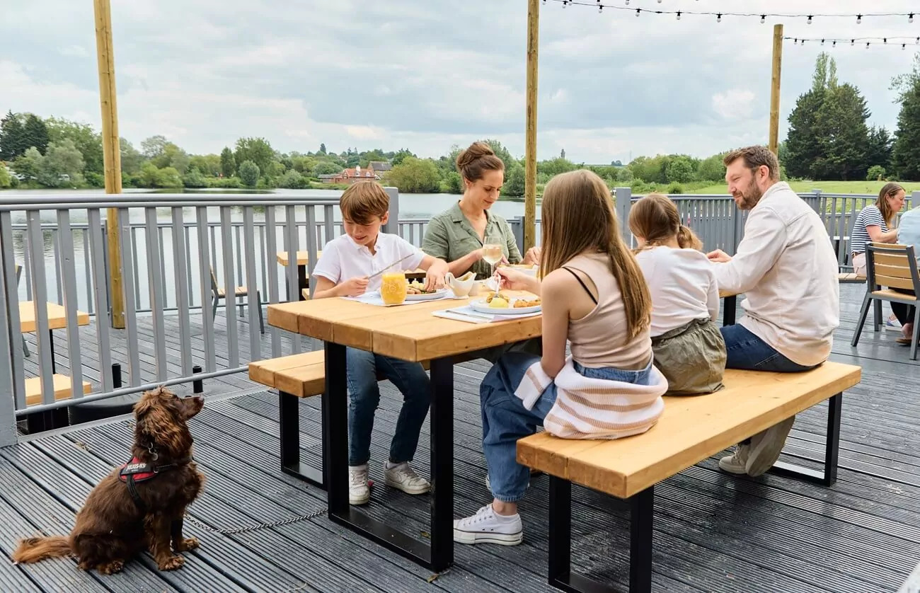 Family enjoying a meal together at a lakeside outdoor dining area, with a brown dog sitting nearby on the deck at a pet-friendly holiday park.