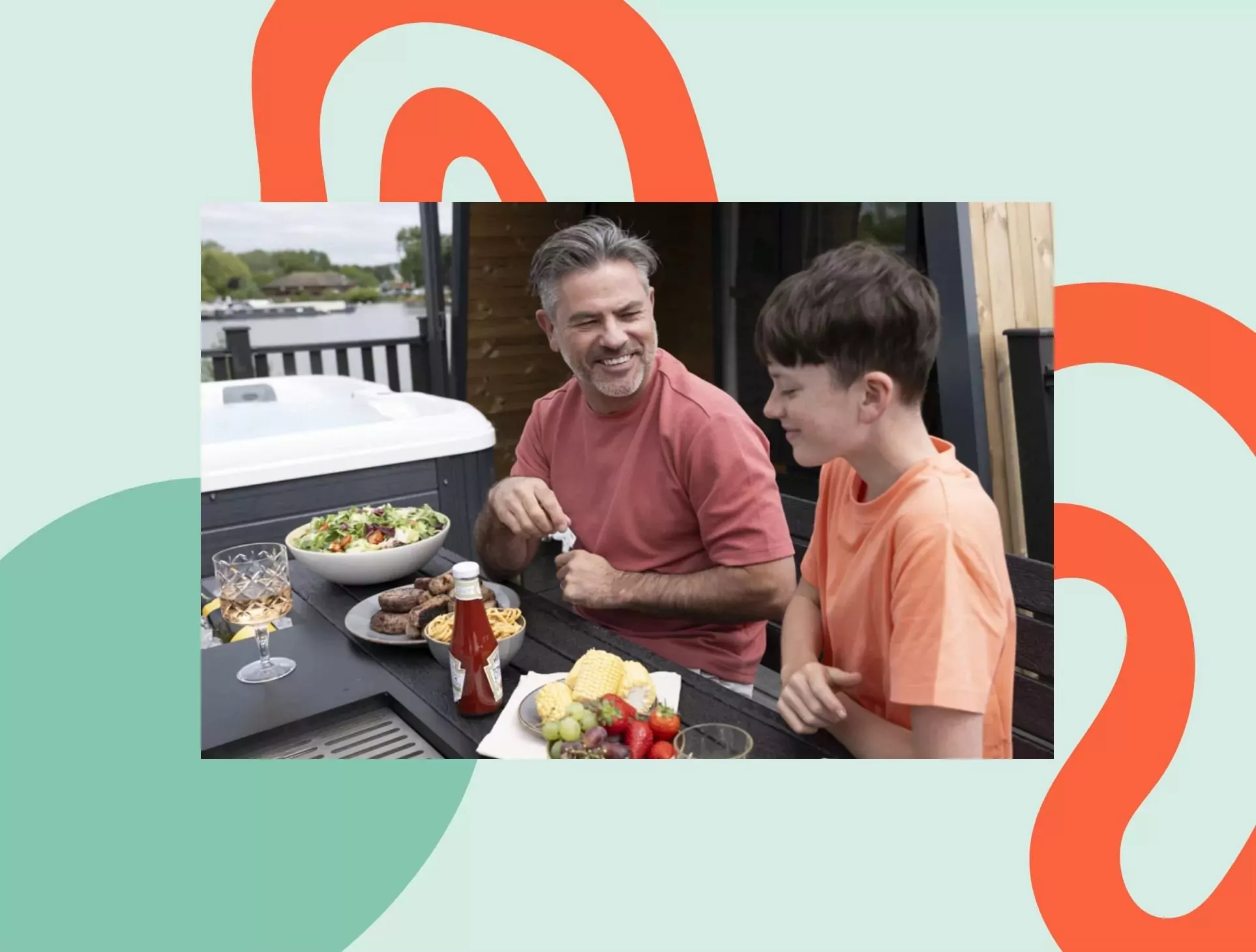 Father and son enjoying an outdoor meal together on a lodge decking, with salad, grilled food, and fruit laid out beside a hot tub at a lakeside holiday park.