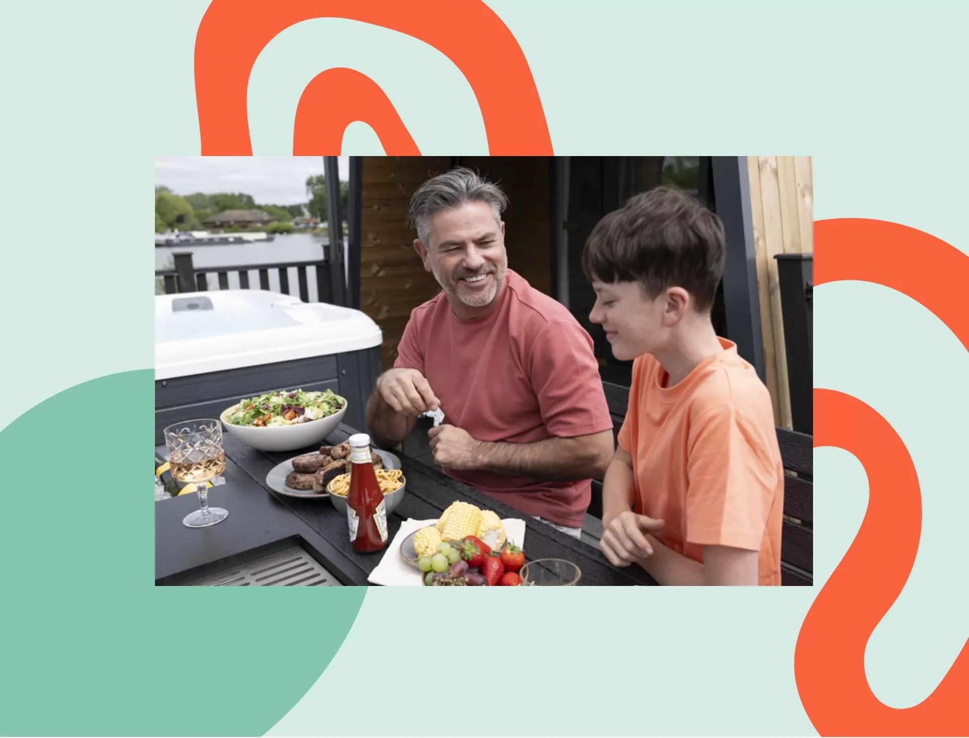 Father and son enjoying an outdoor meal together on a lodge decking, with salad, grilled food, and fruit laid out beside a hot tub at a lakeside holiday park.