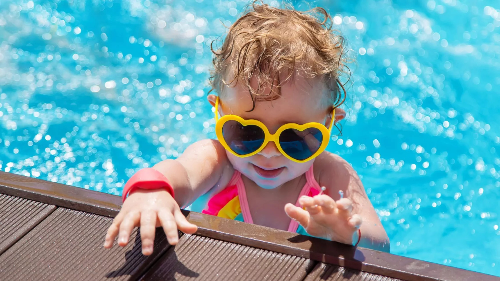 Cute toddler wearing heart-shaped sunglasses climbing out of a swimming pool in bright summer swimwear, enjoying a sunny holiday park day.