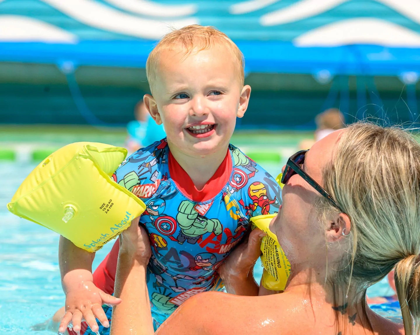 Happy toddler wearing armbands being lifted in a swimming pool with a parent at Meadow Bay Villages