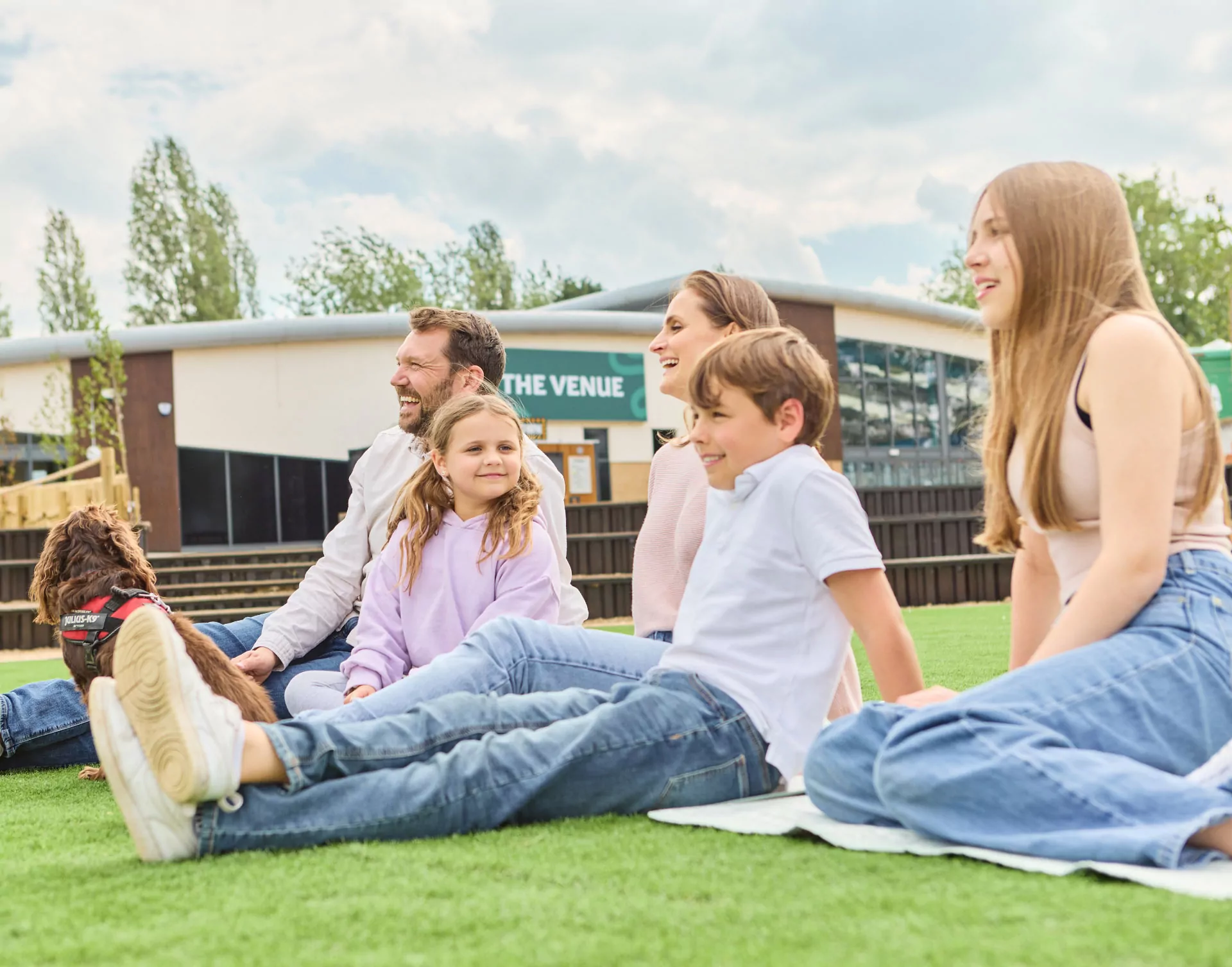 Family relaxing together on green grass outdoors at a holiday park with children and a dog, Meadow Bay Villages