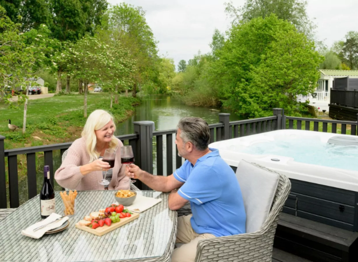 Couple enjoying drinks together on a lodge decking with private hot tub beside a peaceful riverside setting, Meadow Bay Villages