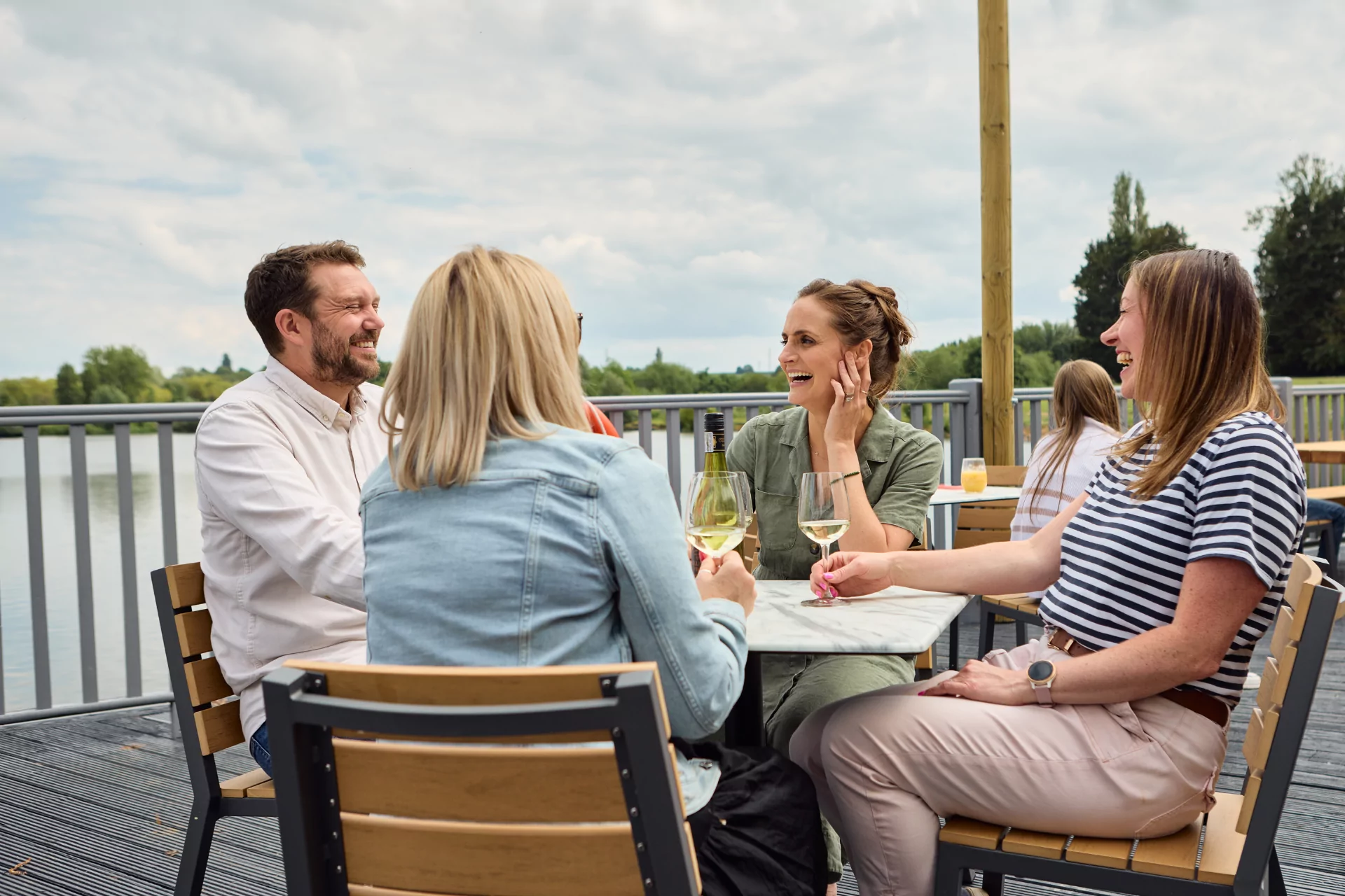 Group of friends enjoying drinks and laughter on a lakeside deck at an outdoor restaurant.