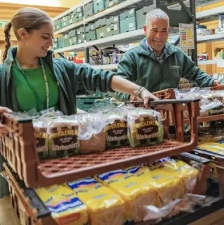 Two volunteers smiling while sorting and stacking loaves of Warburtons bread on trays in a food bank storage area.