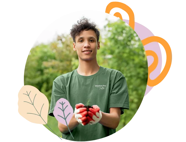Young man wearing a Meadow Bay t-shirt holding a seedling in gardening gloves, smiling with trees in background.
