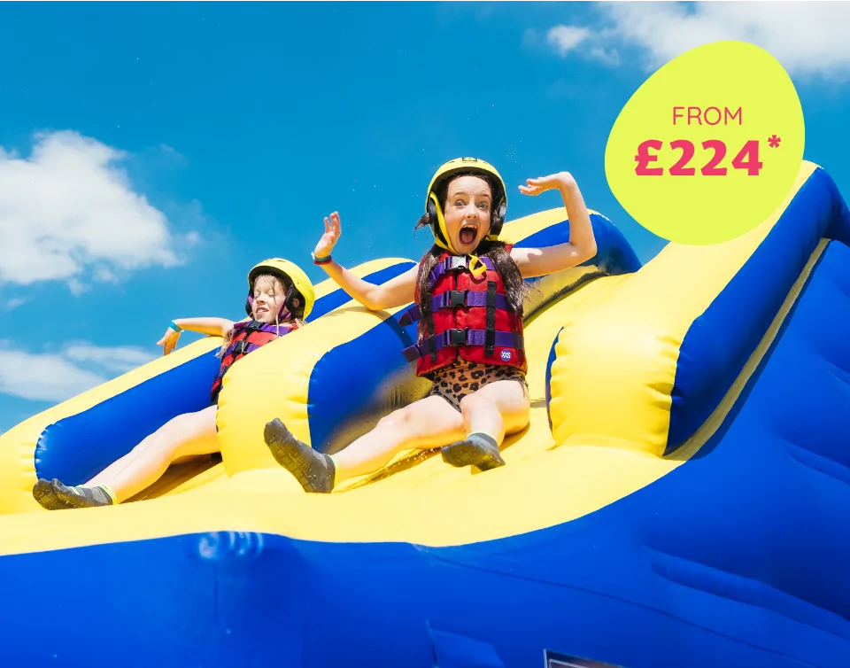 Two excited children sliding down a large inflatable water slide in life jackets on a sunny day, with price promotion from £224.