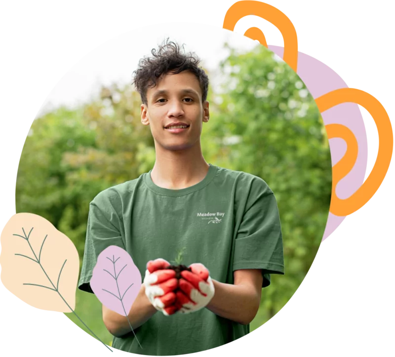Young man wearing a Meadow Bay t-shirt holding a seedling in gardening gloves, smiling with trees in background.