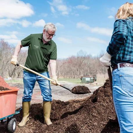 Two people working outdoors, shoveling mulch from a large pile into a wheelbarrow on a sunny day, with trees and open space in the background.