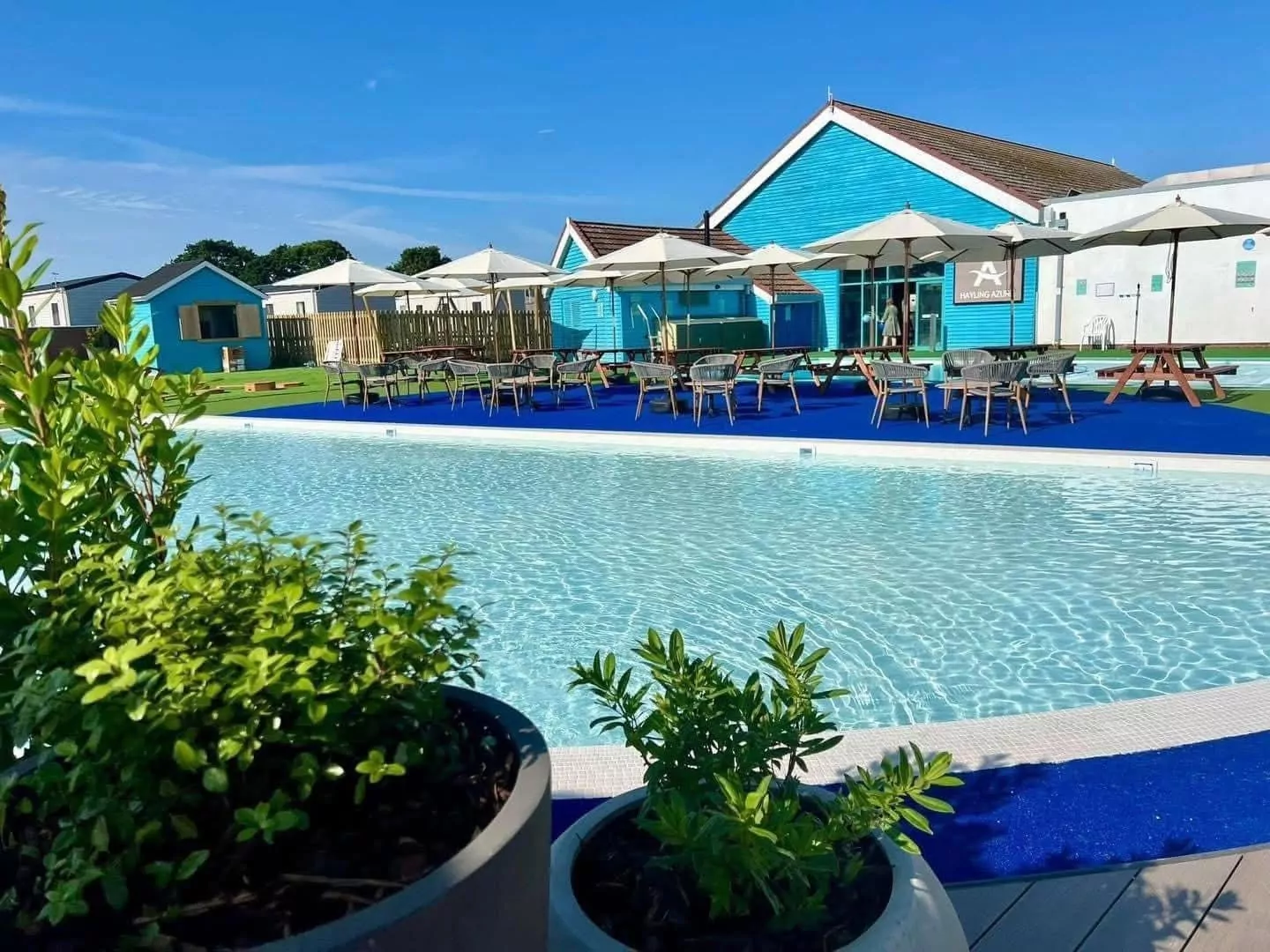 Sunny day at Meadow Bay Villages outdoor pool, with sun loungers, parasols, and blue holiday cabins.