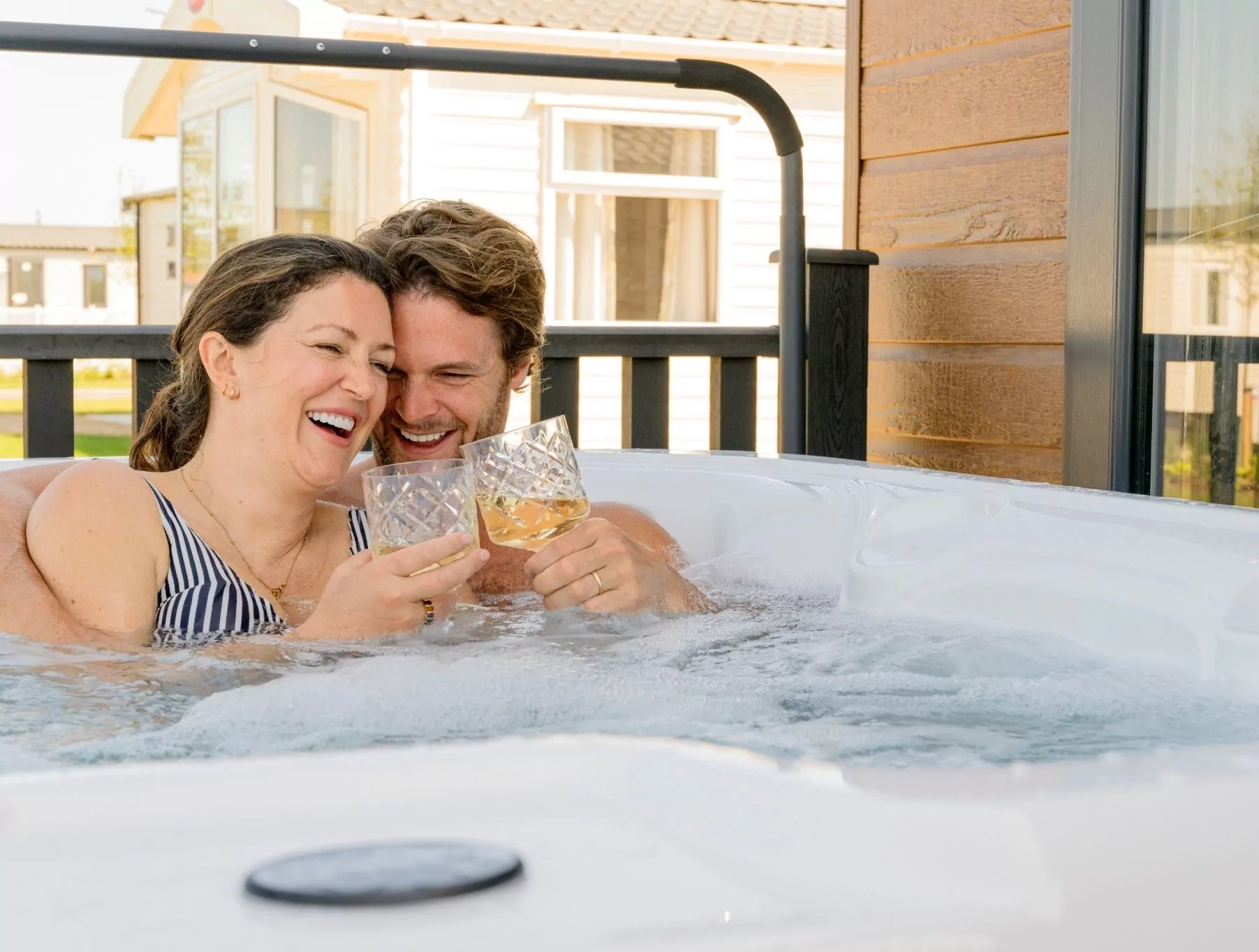 Couple relaxing in a hot tub, smiling and clinking glasses, with holiday lodges in the background.