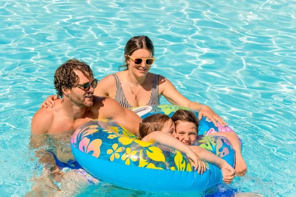 Happy family of four enjoying time together in a bright blue outdoor swimming pool with colourful inflatable rings.