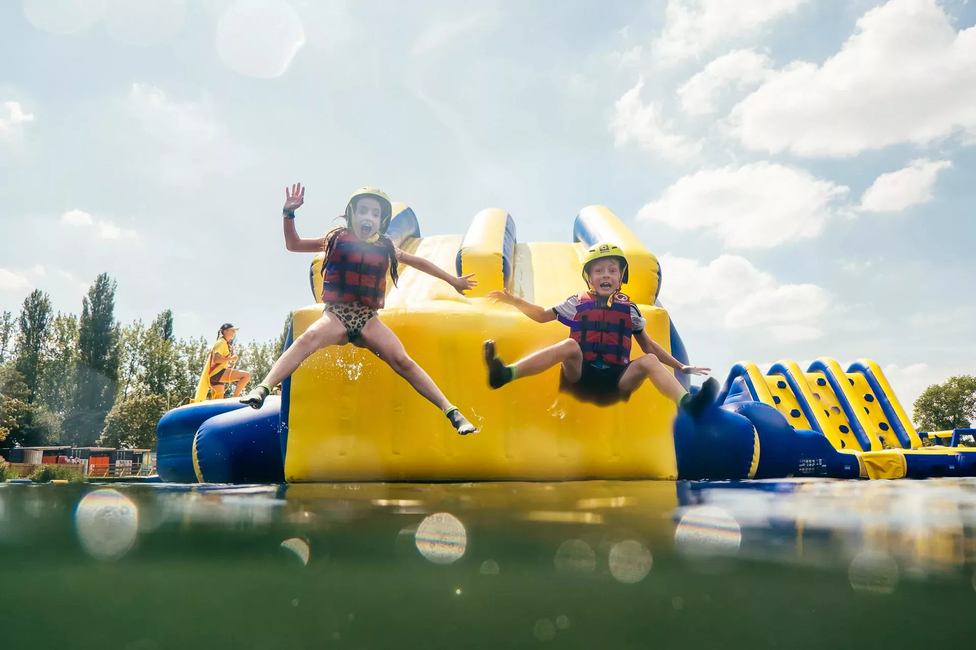 Two excited children in life jackets and helmets jumping into the water from a giant inflatable aqua park obstacle course on a sunny day.