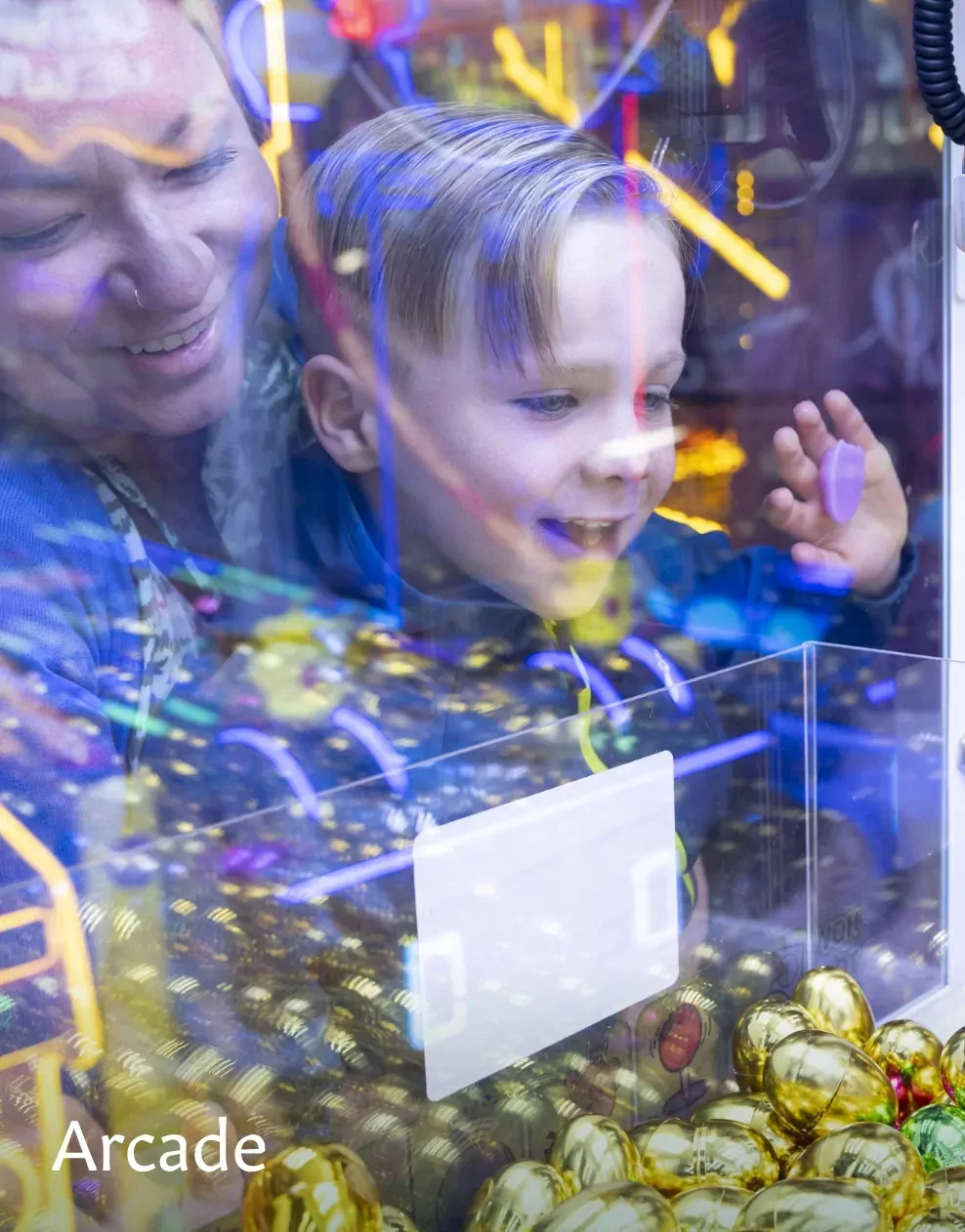 A smiling child playing a claw machine game in the arcade with a parent at Meadow Bay Villages.