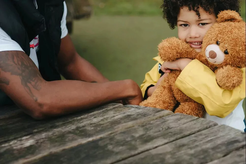 Young child smiling while hugging a teddy bear at a wooden picnic table, wearing a yellow coat during a cuddly craft activity at the holiday park.