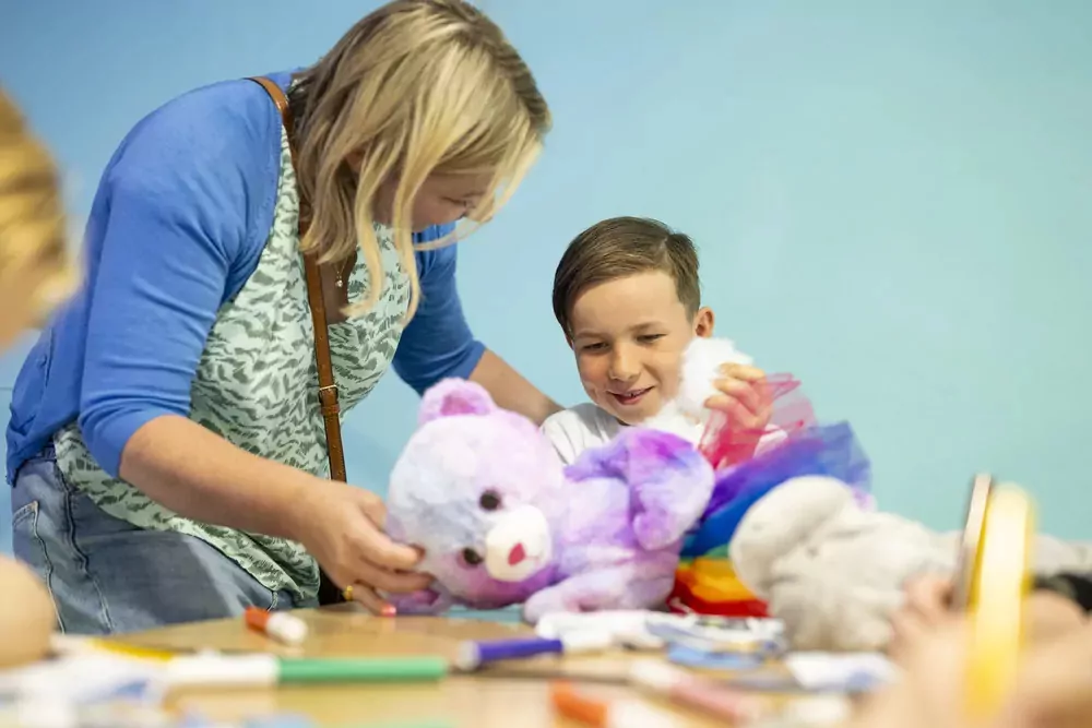 A smiling child creating a colourful stuffed bear with the help of an adult at the Build-A-Bear workshop in Meadow Bay Villages.