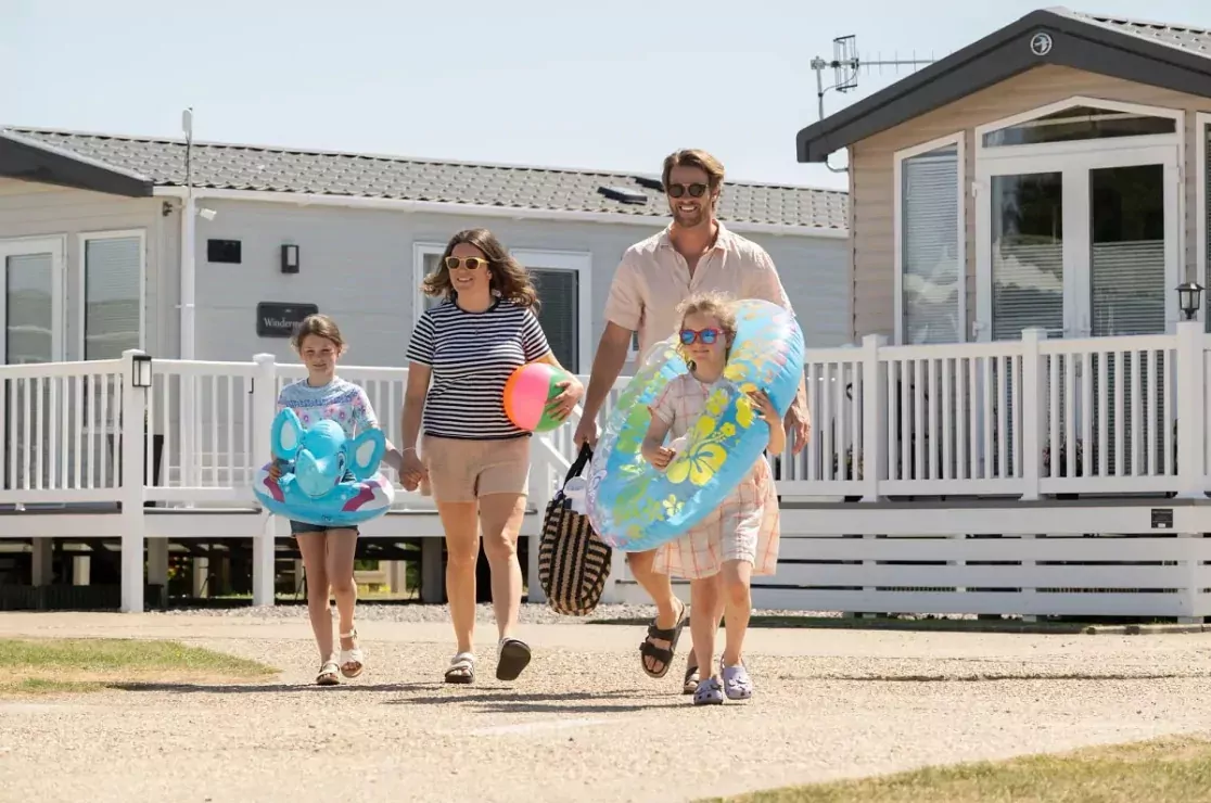 Smiling family walking from a caravan holiday home carrying beach toys and floaties, heading out for a fun day.