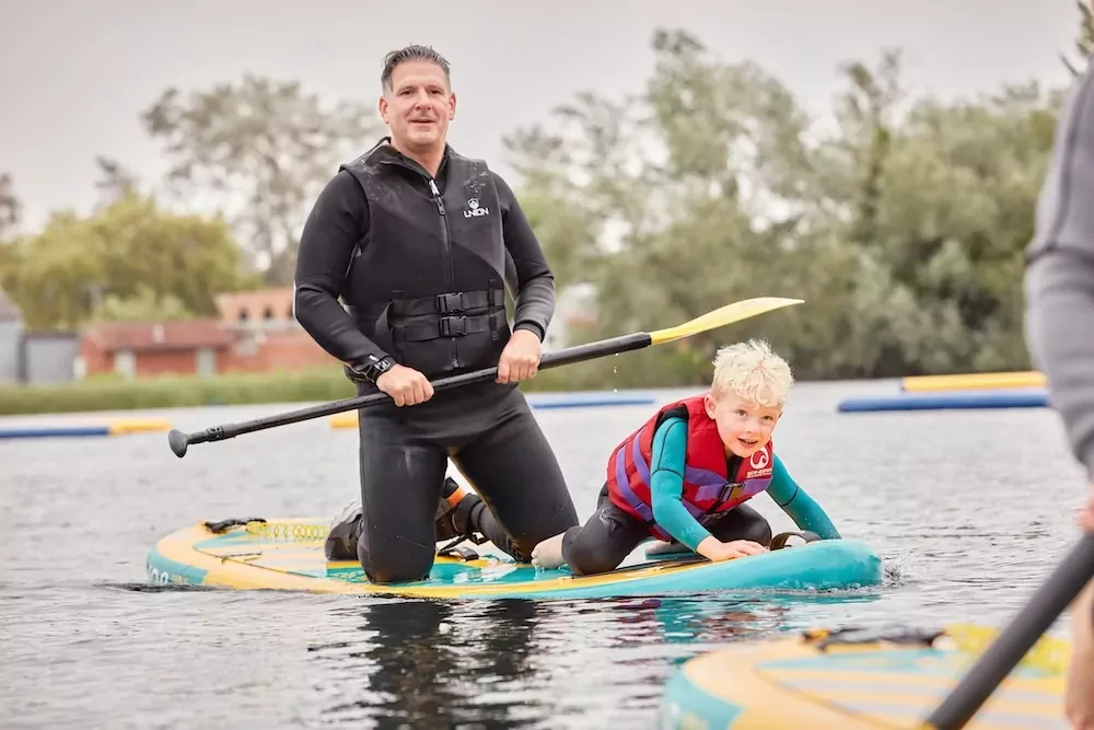 Father and young son wearing wetsuits and life jackets enjoying a paddleboarding session on a calm lake at a holiday park.