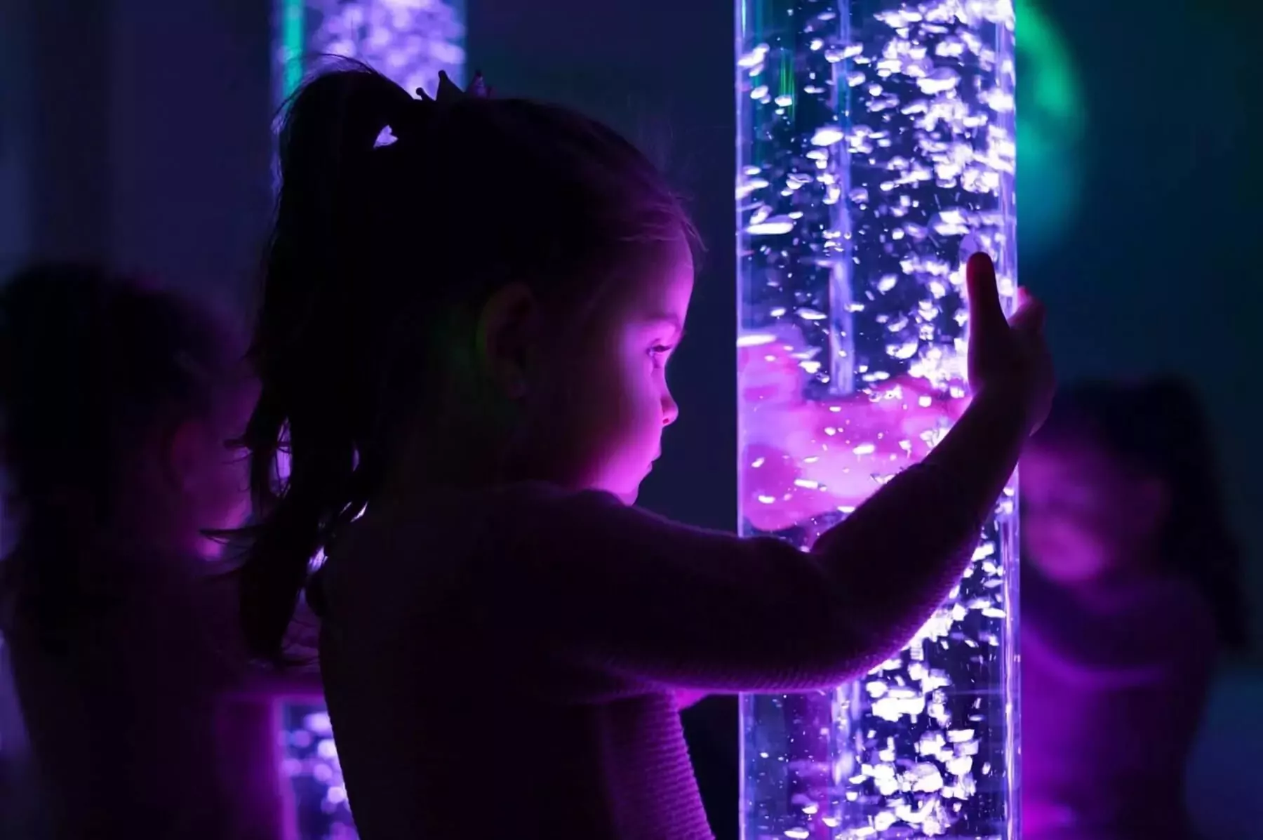 Young girl interacting with a glowing bubble tube in a dark, calming sensory room, lit with purple and blue lights for a relaxing experience.