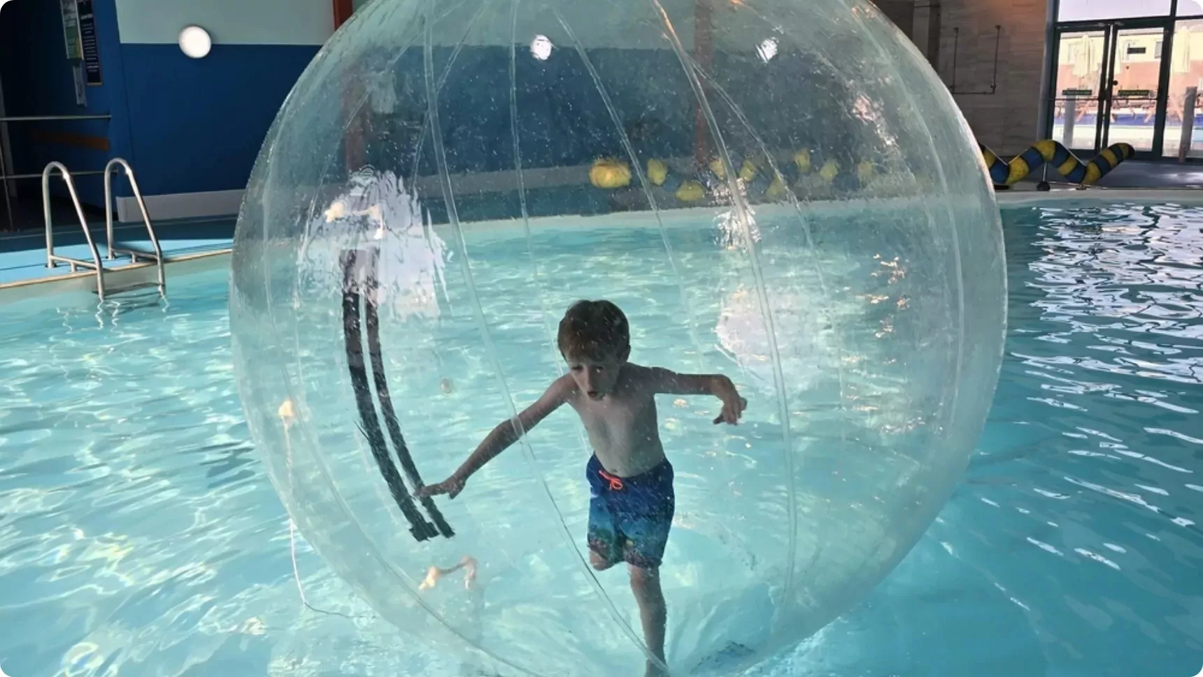 Young boy inside a giant inflatable zorb ball floating on the surface of an indoor swimming pool at a holiday park.