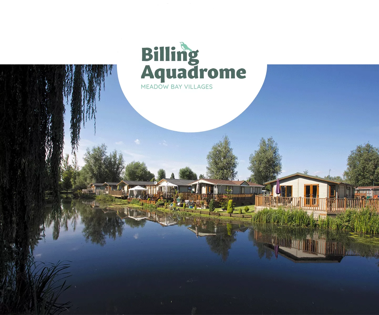 Row of holiday lodges beside a tranquil lake with trees and clear reflections under a blue sky at Billing Aquadrome, Meadow Bay Villages.