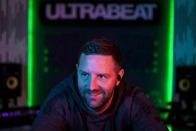A man in a hoodie smiles with "ULTRABEAT" sign behind him.