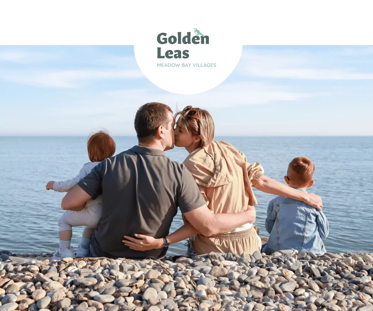 Young family sitting on a pebble beach, parents kissing while kids look out over the calm sea at Golden Leas holiday village.