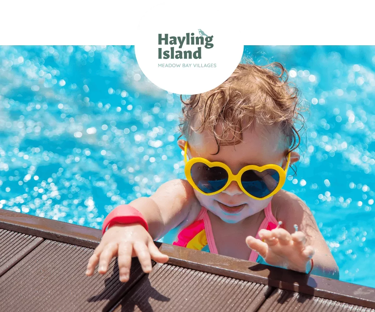 Young child in heart-shaped sunglasses and a pink swimsuit playing by the edge of a bright blue swimming pool at Hayling Island.