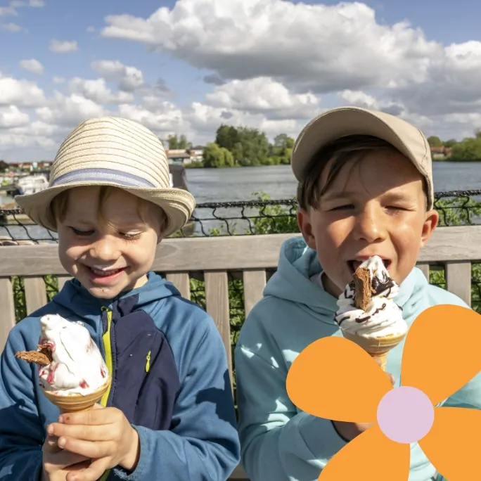 Two young boys sitting on a bench by a lake, smiling and enjoying ice cream cones on a sunny day.