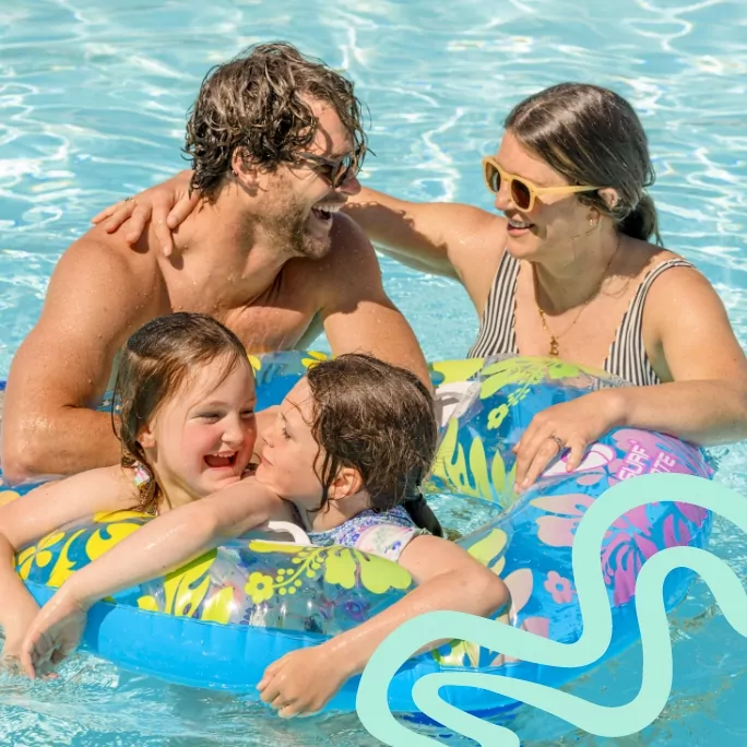 Family of four laughing together in a swimming pool, with two children in colourful inflatable rings.