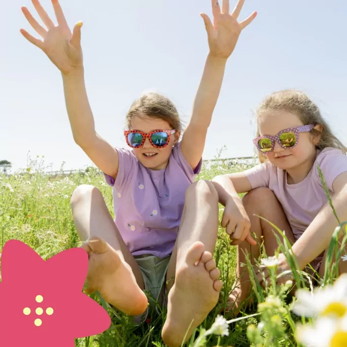 Two young girls sitting in a wildflower field on a sunny day, smiling and wearing colourful sunglasses.